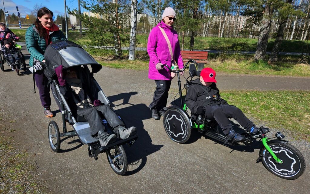 On aurinkoinen sää. Kaksi aikuista ja kaksi lasta kulkevat hiekkatiellä. Toinen lapsista istuu maastorattaissa, joissa on syvä istuin, kasvoja varjostava kuomu ja kolme suurta rangasta. Toinen aikuisista työntää rattaita takaapäin. Toinen lapsi istuu kolmipyörässä, jossa istuinasento on taaksepäin nojaava. Jalat ovat polkimilla, ja ne osoittavat eteenpäin. Pyörän sarvet ovat sivuilla pystysuuntaisesti. Takana kävelevä aikuinen ohjaa pyörää toisista, takana olevista sarvista.
