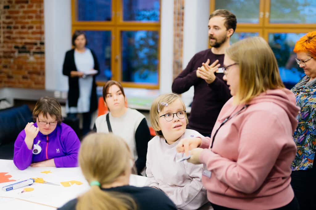 Ihmisiä keskustelee tilaisuudessa. Osa istuu pöydän ääressä, osa seisoo ympärillä. Taustalla tulkki tulkkaa keskustelua.
