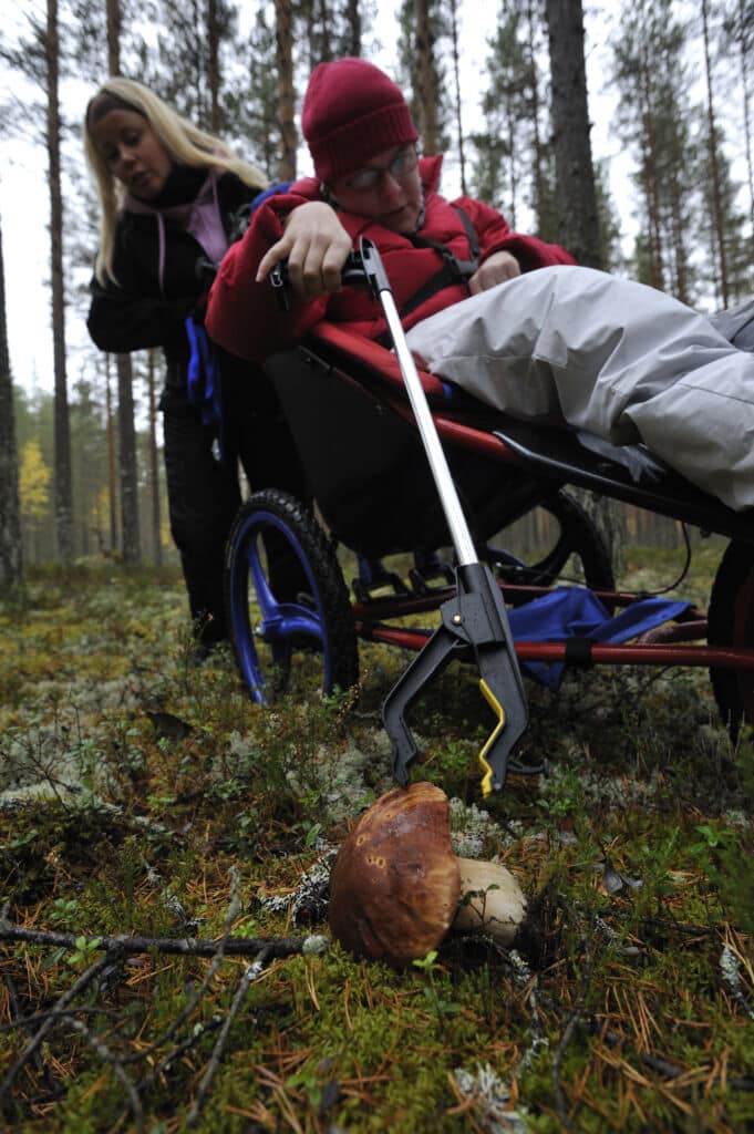 Maastpyörätuolissa istuva ihminen poimii sienen maasto varrellisten pihtien avulla. Pihdit sulkeutuvat käsikahvasta purstimalla.