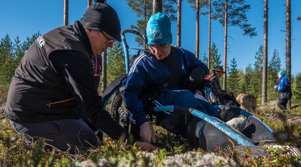 Kaksi miestä poimii sormin marjoja sammalmättäällä. Toinen on kyykyssä ja toinen istuu matalassa maastopyörätuolissa.