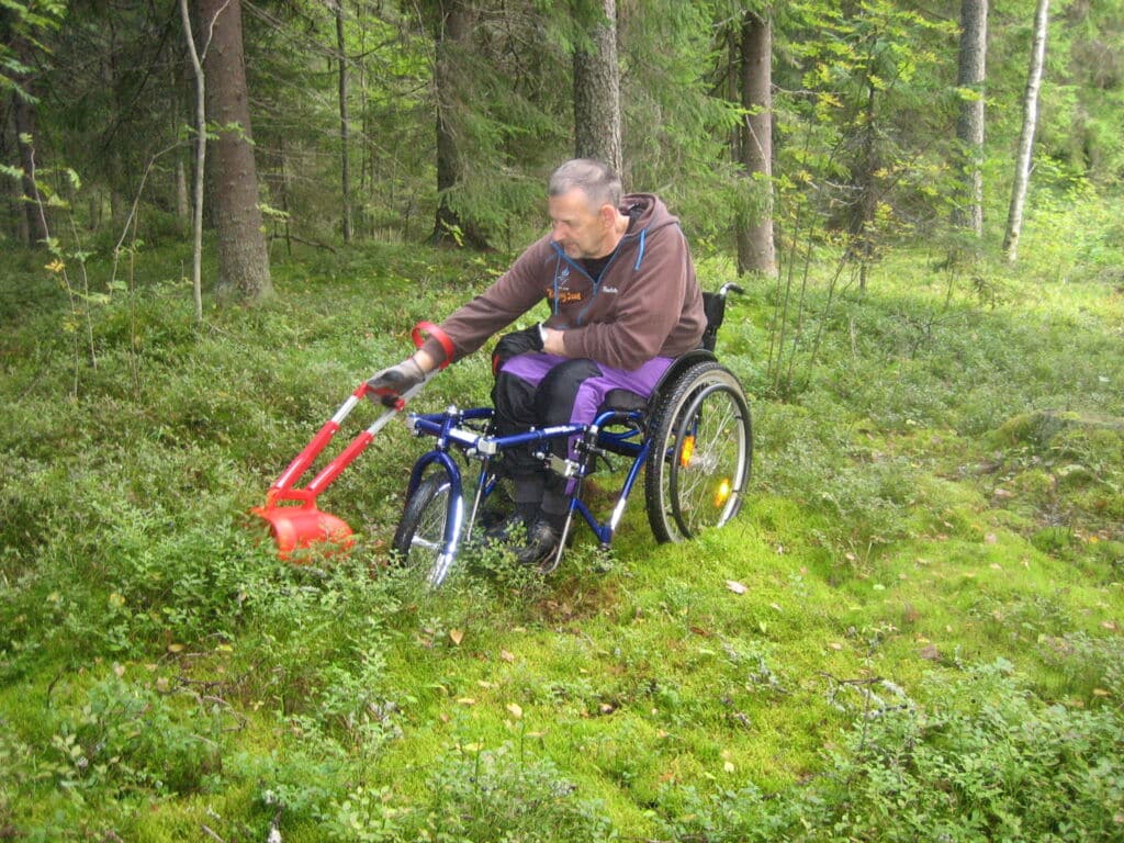 Mies istuu pyörätuolissa, johon on kiinnitetty isorenkainen nokkapyörä. Miehellä on kädessään pitkävartinen marjapoimuri, jolla hän poimii marjoja pyörätuolista käsin. Poimurin varressa on pystykahva ja kyynärtuki.