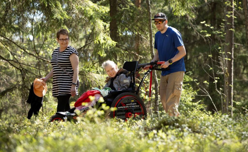 Kaksi aikuista ja yksi lapsi liikkuvat aurinkoisessa metsässä. Lapsi istuu maastopyörätuolissa, jossa on takana kaksi isoa rengasta ja edessä yksi pienempi rengas.