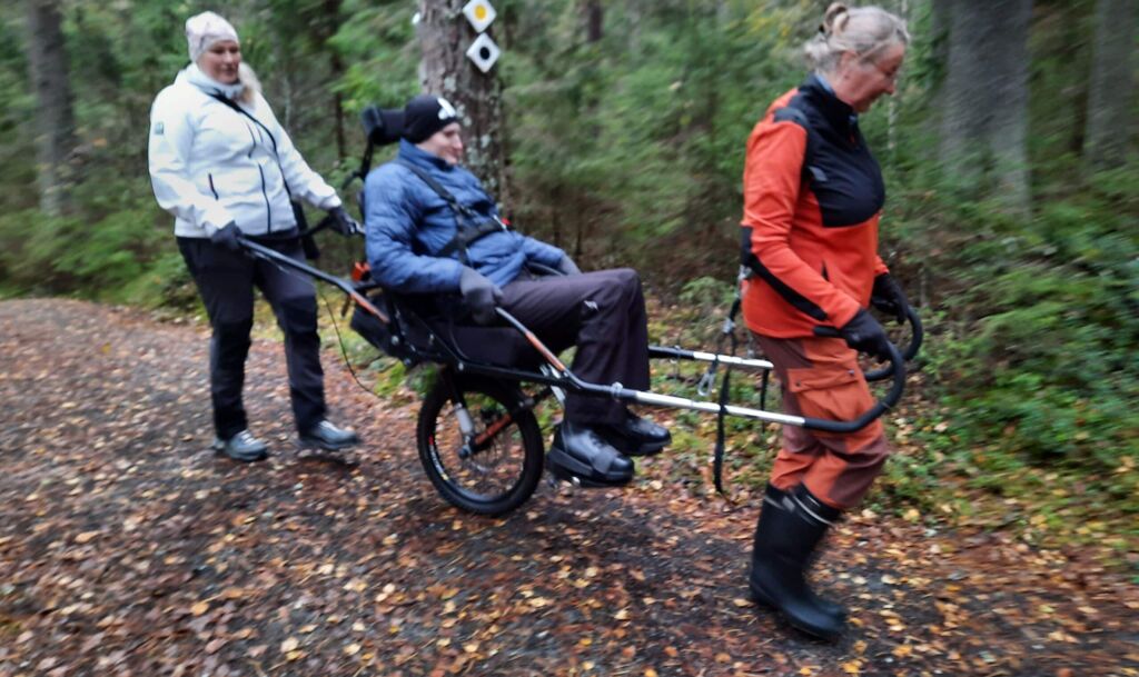 Mies istuu yksipyöräisessä maastokärryssä. Yksi avustaja vetää edessä olevista vetoaisoista ja toinen avustaja työntää takana olevista työntöaisoista. Aisat ovat metalliset.