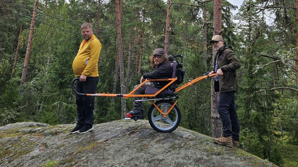 Mies istuu yksipyöräisessä maastokärryssä kalliolla. Toinen mies seisoo edessä pitäen kiinni kärryn vetoaisoista ja kolmas mies työntää takaa työntöaisoista.
