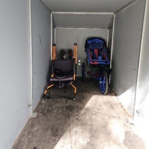 Sininen Kangoo-monitoimiratas peräkärryn oikeassa takanurkassa. Selkänoja on vasten peräkärryn takaseinää. Kangoon vieressä, peräkärryn vasemmassa takanurkassa on oranssi-musta Joelette-maastokärry selällään. Vetoaisat on taitettu kokoon kärryn sivuille. Joeletten istuinosa osoittaa koti peräkärryn oviaukkoa.