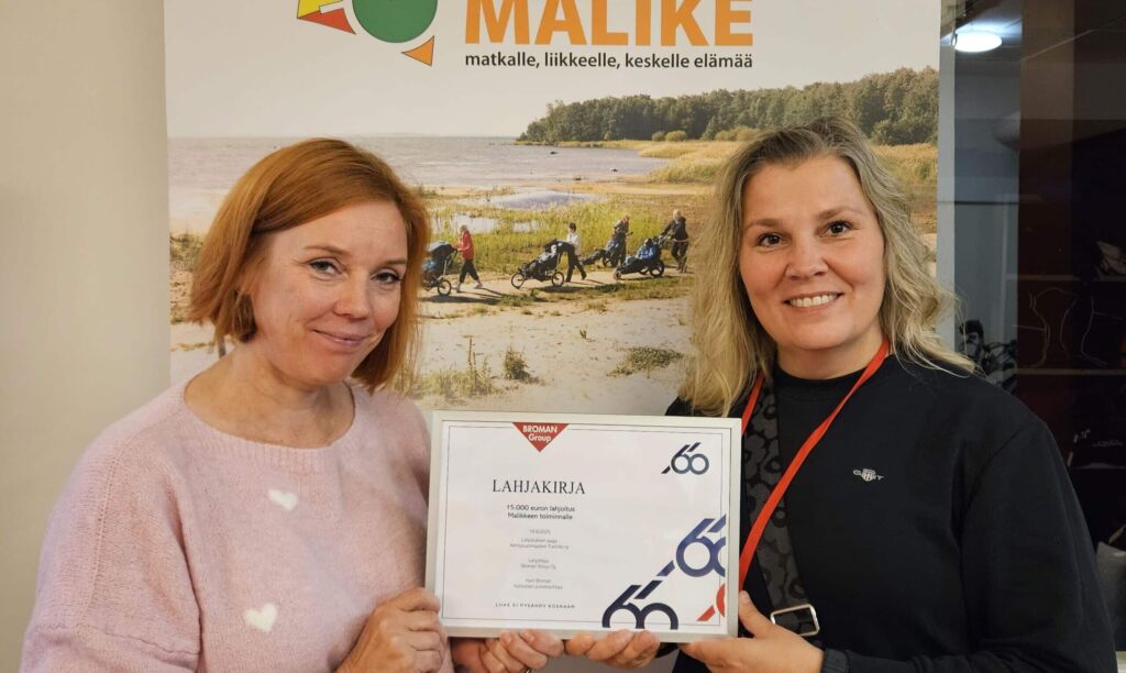 Susanna Tero ja Katja Pohtola poseeraavat Malike-roll-upin edessä pitäen kiinni kehystetystä lahjakirjasta.