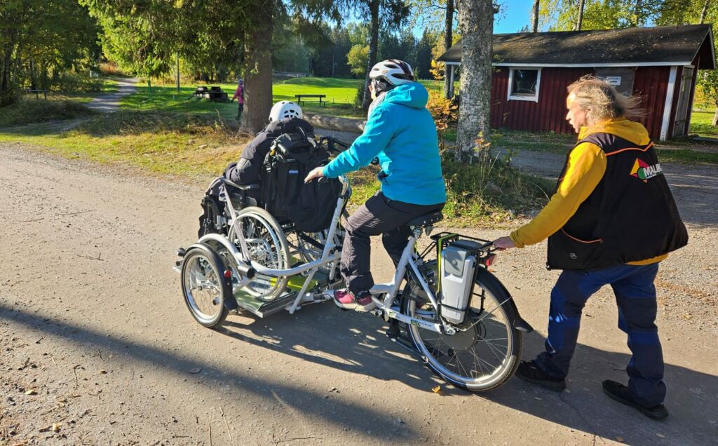 Nainen polkee ja ohjaa kolmpiyörää, jonka etuosassa on lava, jolle on nostettu ihminen manuaalipyörätuolissa. Takana Malikkeen työntekijä työntää vauhtia.